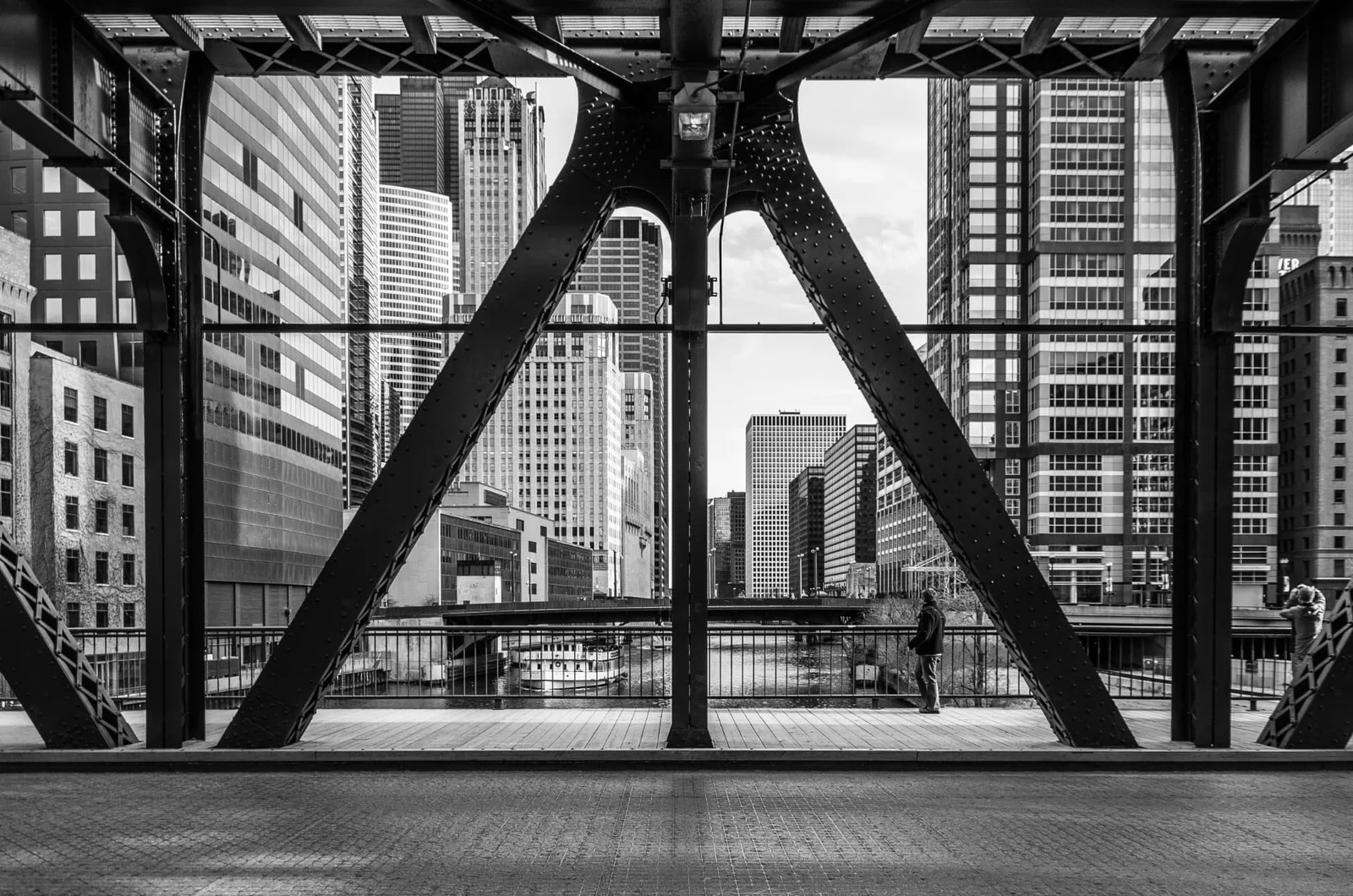 Black-and-white view through the steel trusses of a Chicago river bridge toward downtown towers.